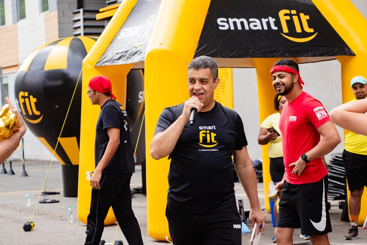 Apresentador ao microfone em frente ao pórtico inflável da Smart Fit, com instrutores ao lado